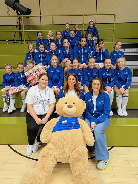 Die Dancers in blauen Trainingsjacken auf einer Hallentribüne, vorne ein großer Teddybär