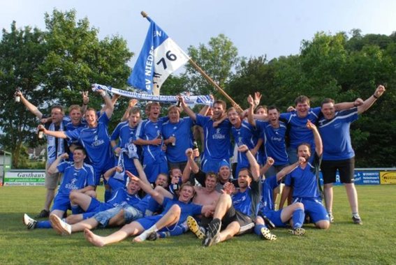 Mannschaft in blauen Trikots feiert auf dem Rasen mit einer Fahne mit der Aufschrift 76