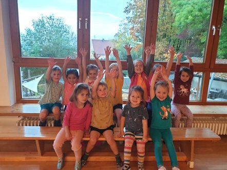 Kindergruppe sitzt mit erhobenen Armen auf einer Bank vor großen Fenstern